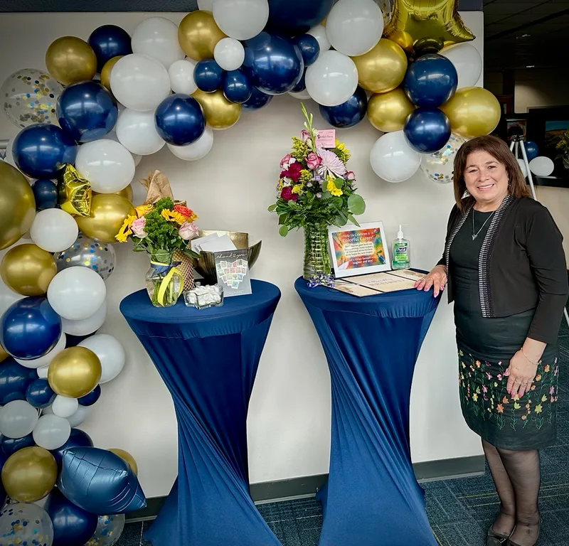 Dora standing beside two tables with flowers under a navy, white and gold balloon arch.