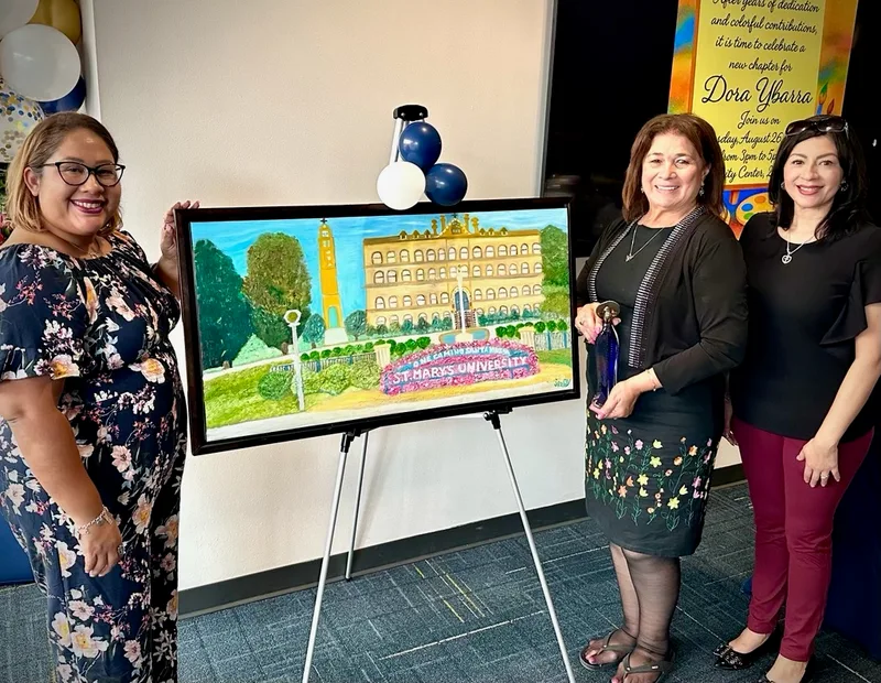 Dora and two friends smiling beside her framed painting of St. Mary’s University on an easel.